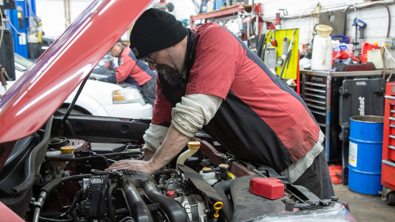 A technician leans over an open vehicle hood to inspect and service components in the engine bay