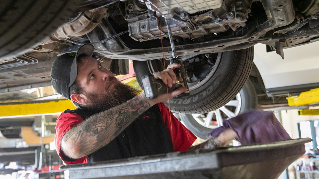 A technician works underneath a lifted vehicle draining fluid into an oil drain pan in a service bay