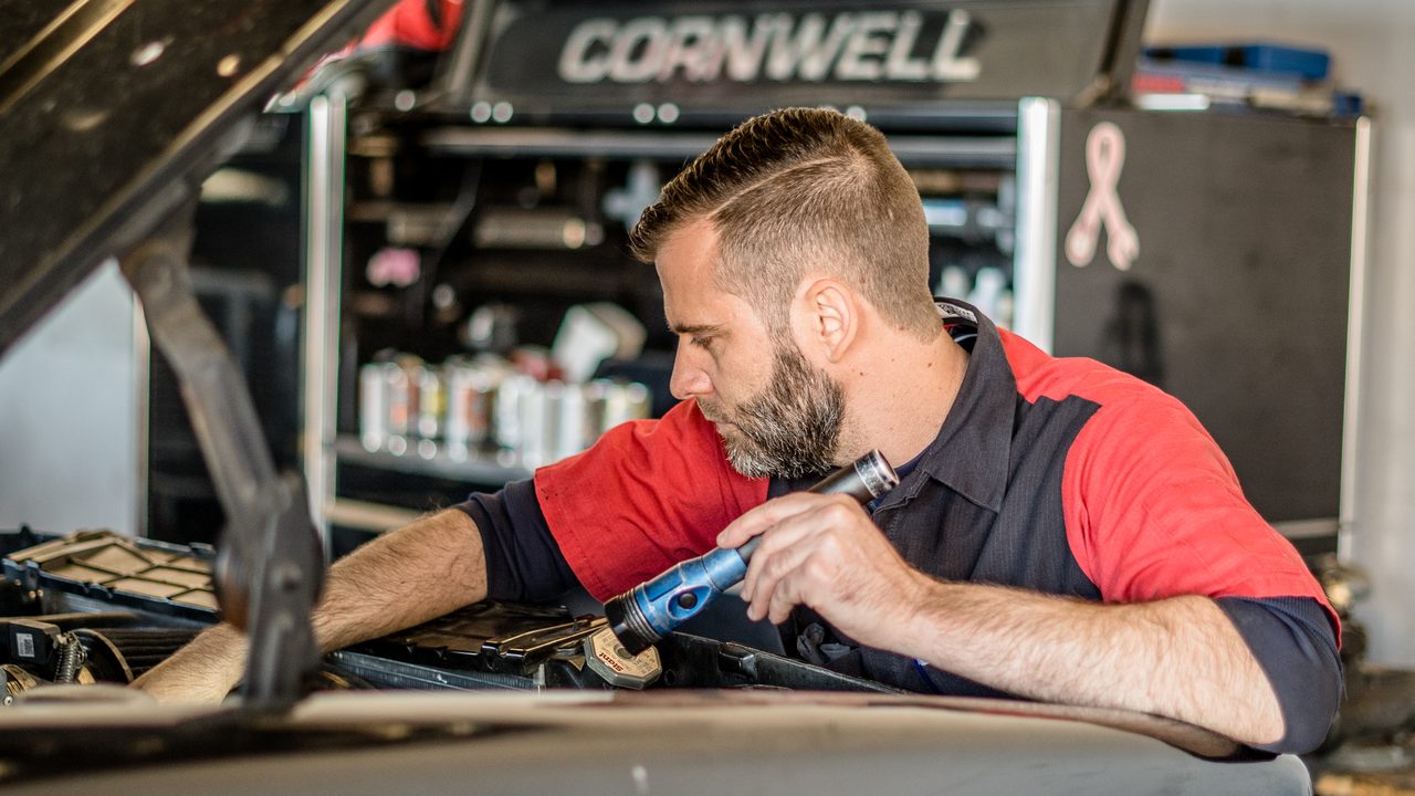 A technician inspects a vehicle's engine bay using a flashlight in an auto repair shop