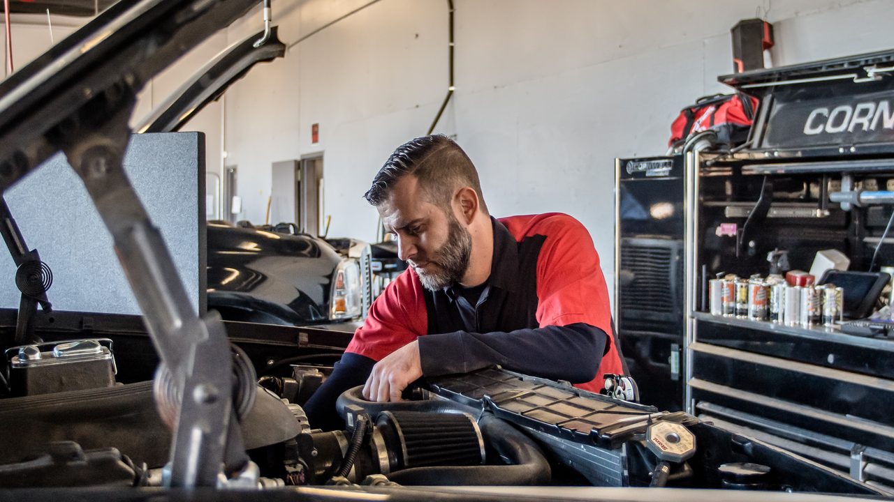 A technician inspects components inside the open engine bay of a car in a service shop