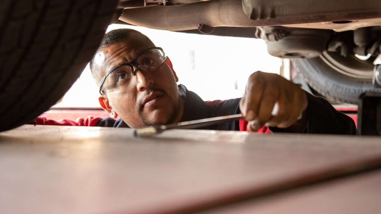 A technician wearing safety glasses inspects and works on the undercarriage of a vehicle on a lift