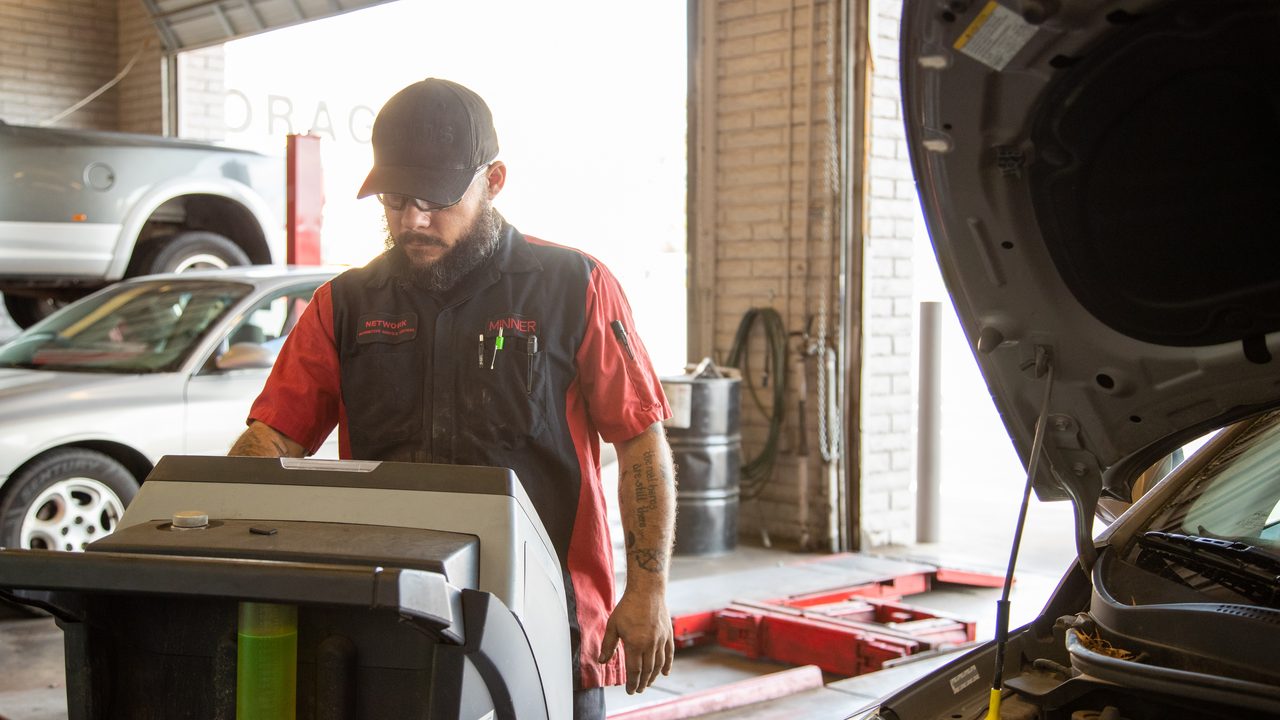 A technician operates an AC service machine beside a vehicle in an auto repair shop bay