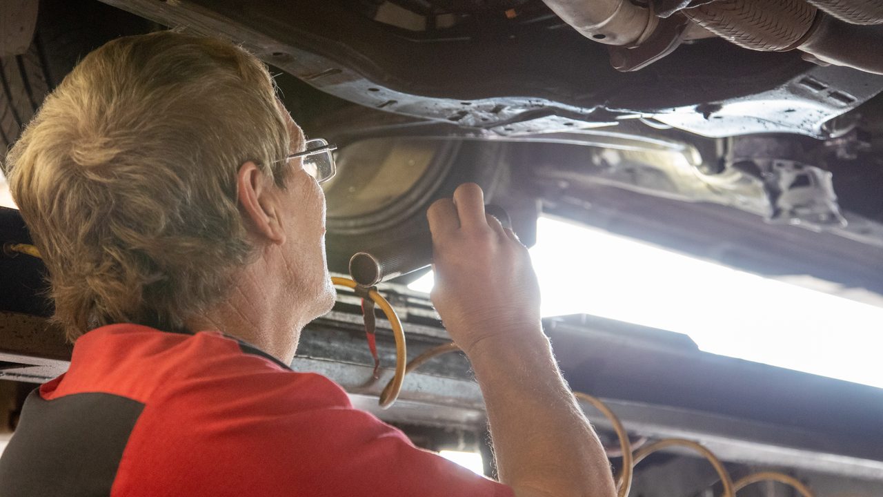 A technician uses a flashlight to inspect the undercarriage and exhaust system of a car on a lift