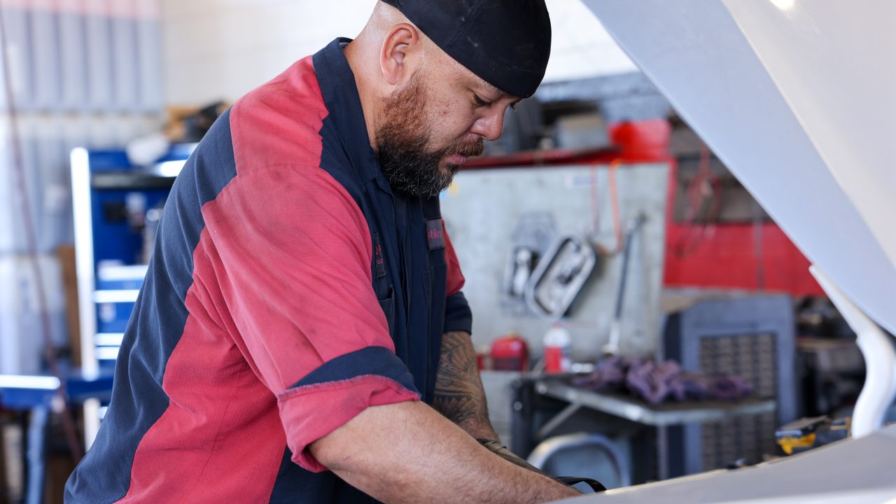A technician works under the open hood of a vehicle inside an auto repair shop
