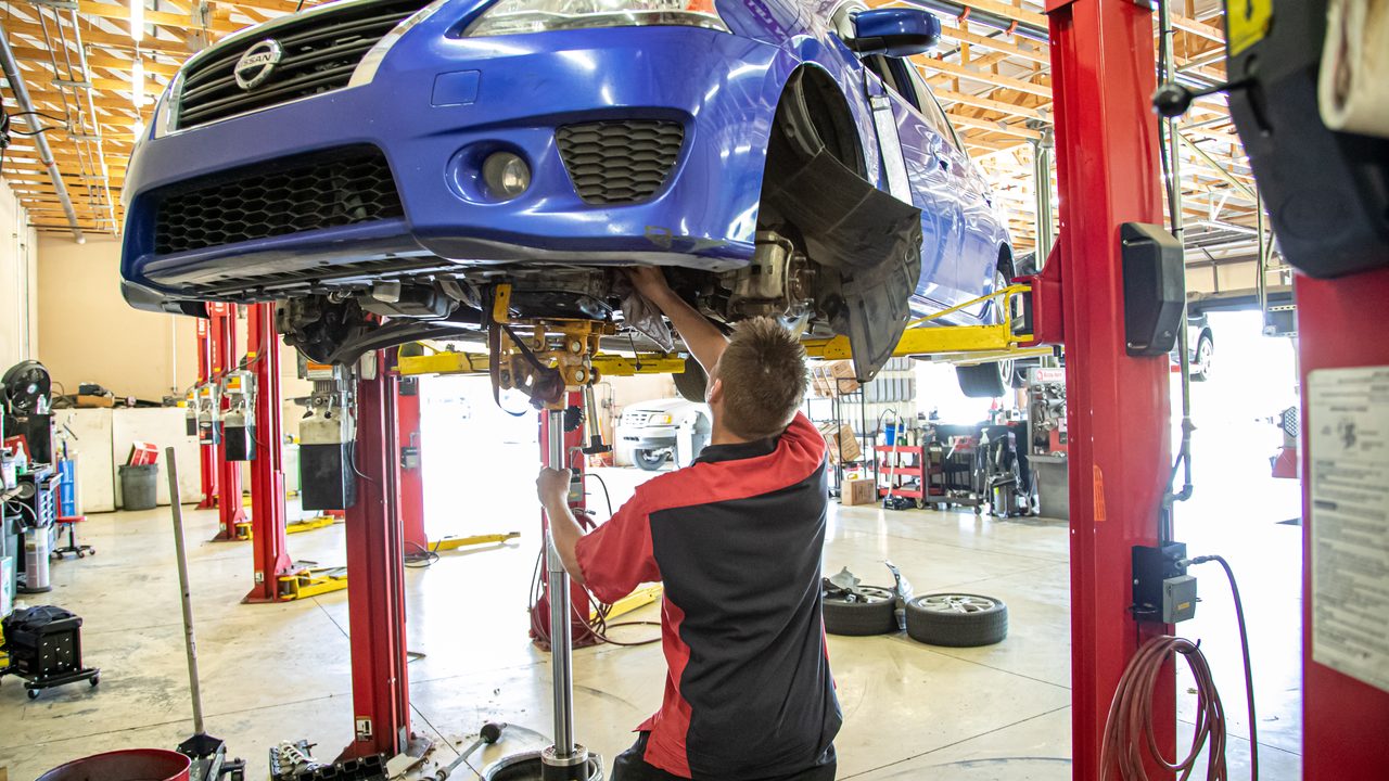 A technician works on the undercarriage of a blue sedan raised on a lift in a service bay