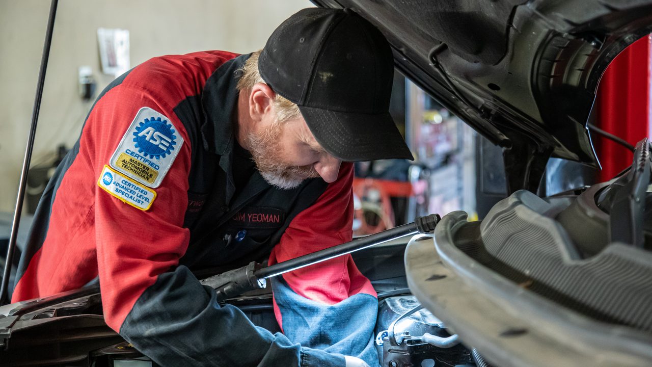 An ASE-certified technician inspects components inside an open engine bay in an auto repair shop