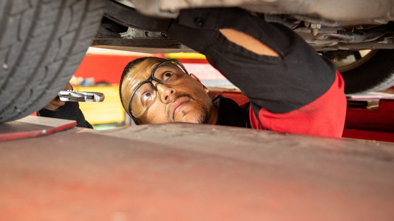 A technician wearing safety glasses inspects the undercarriage and suspension of a vehicle on a lift