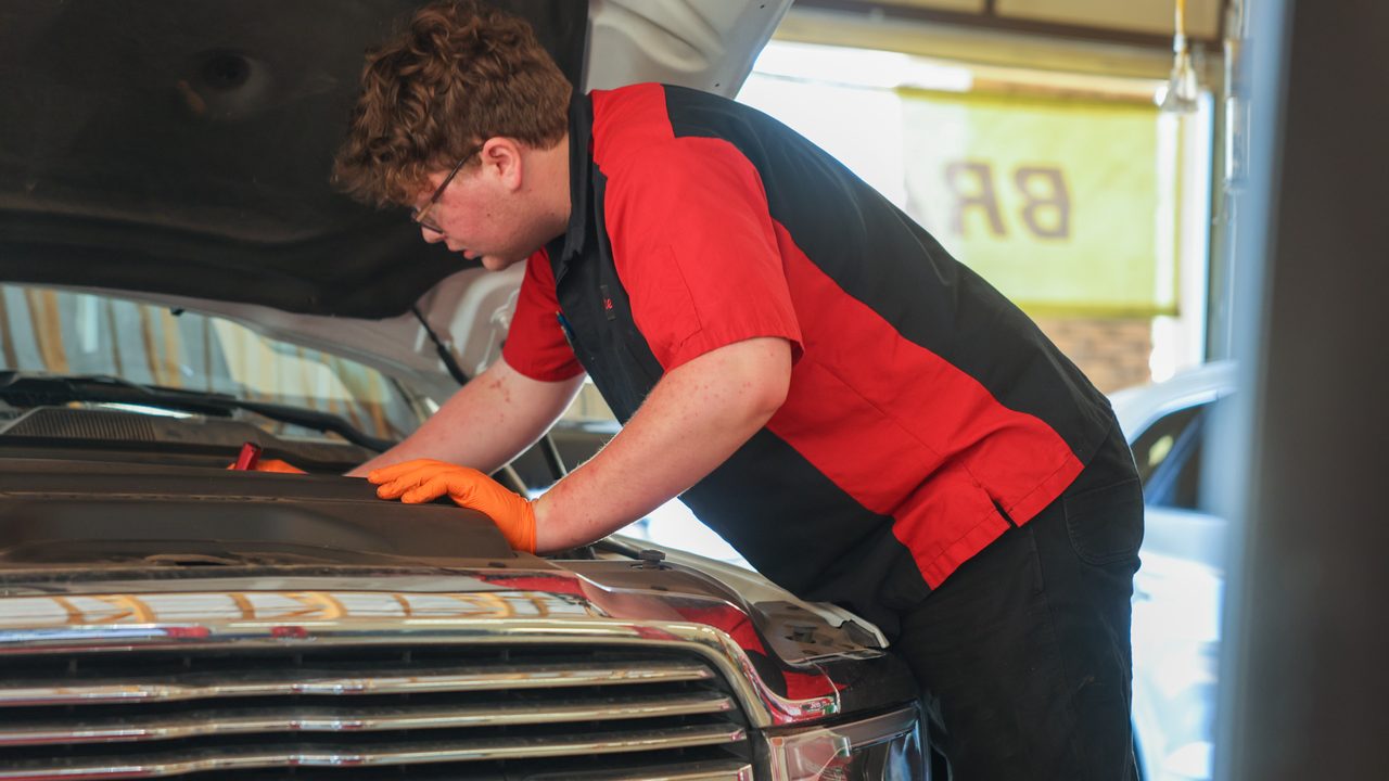 A technician wearing orange gloves inspects the engine bay of a RAM pickup truck inside a service bay