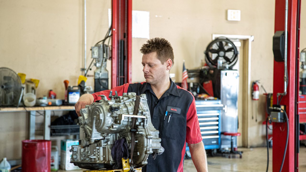 A technician inspects a removed transmission mounted on a stand inside a service bay