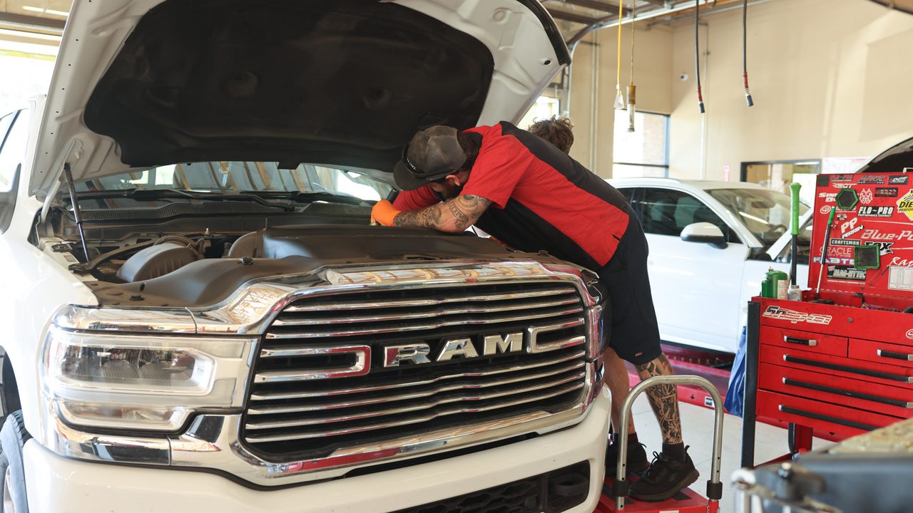 A technician leans into the engine bay of a white pickup truck inside an auto repair shop