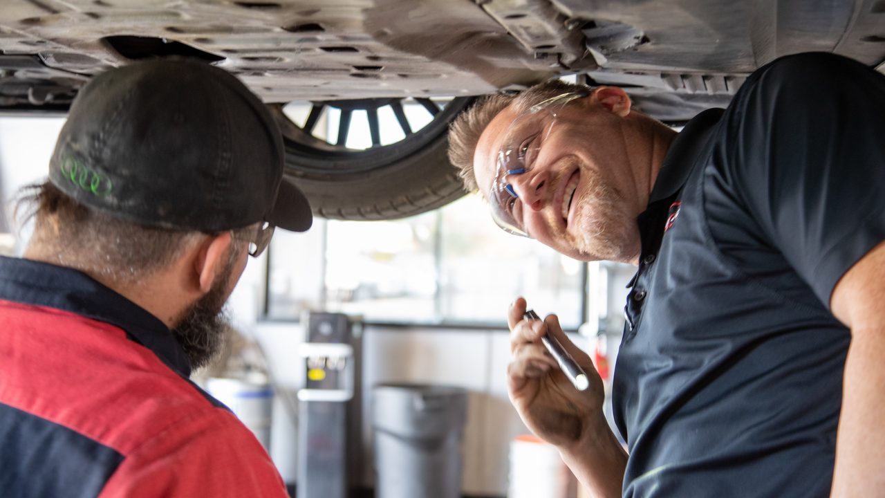 Two technicians inspect the undercarriage of a raised vehicle using a flashlight in a service bay