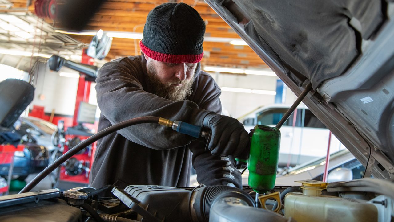A technician uses a pneumatic tool to work inside the vehicle's engine bay in an auto repair shop