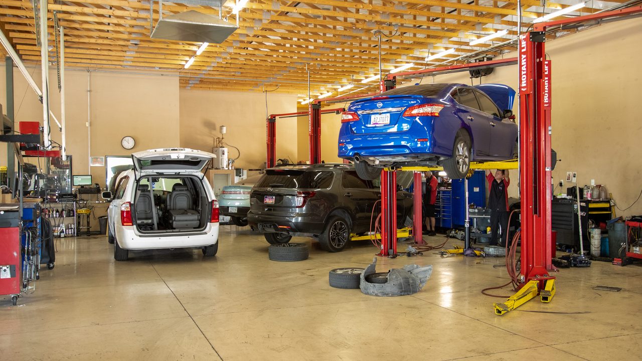 A technician works beneath a blue sedan raised on a lift inside a busy automotive repair shop