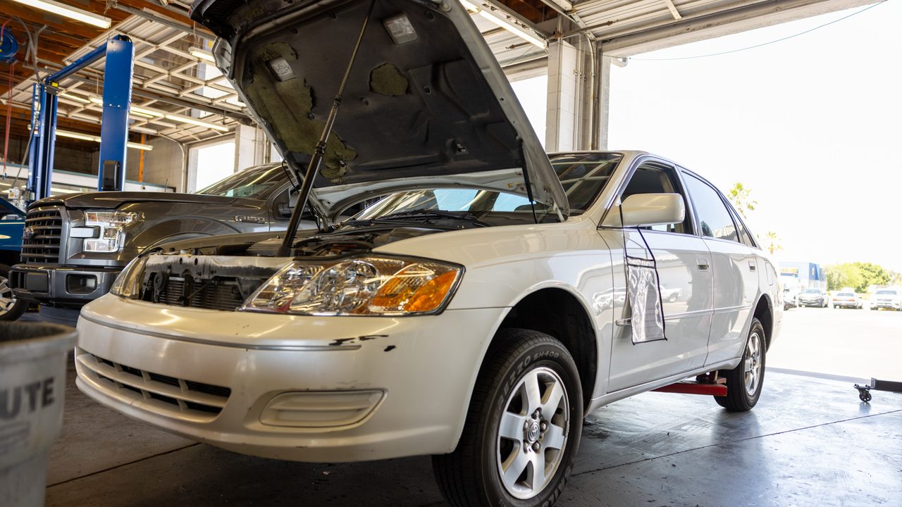 A white sedan with its hood open sits elevated inside a brightly lit auto repair service bay