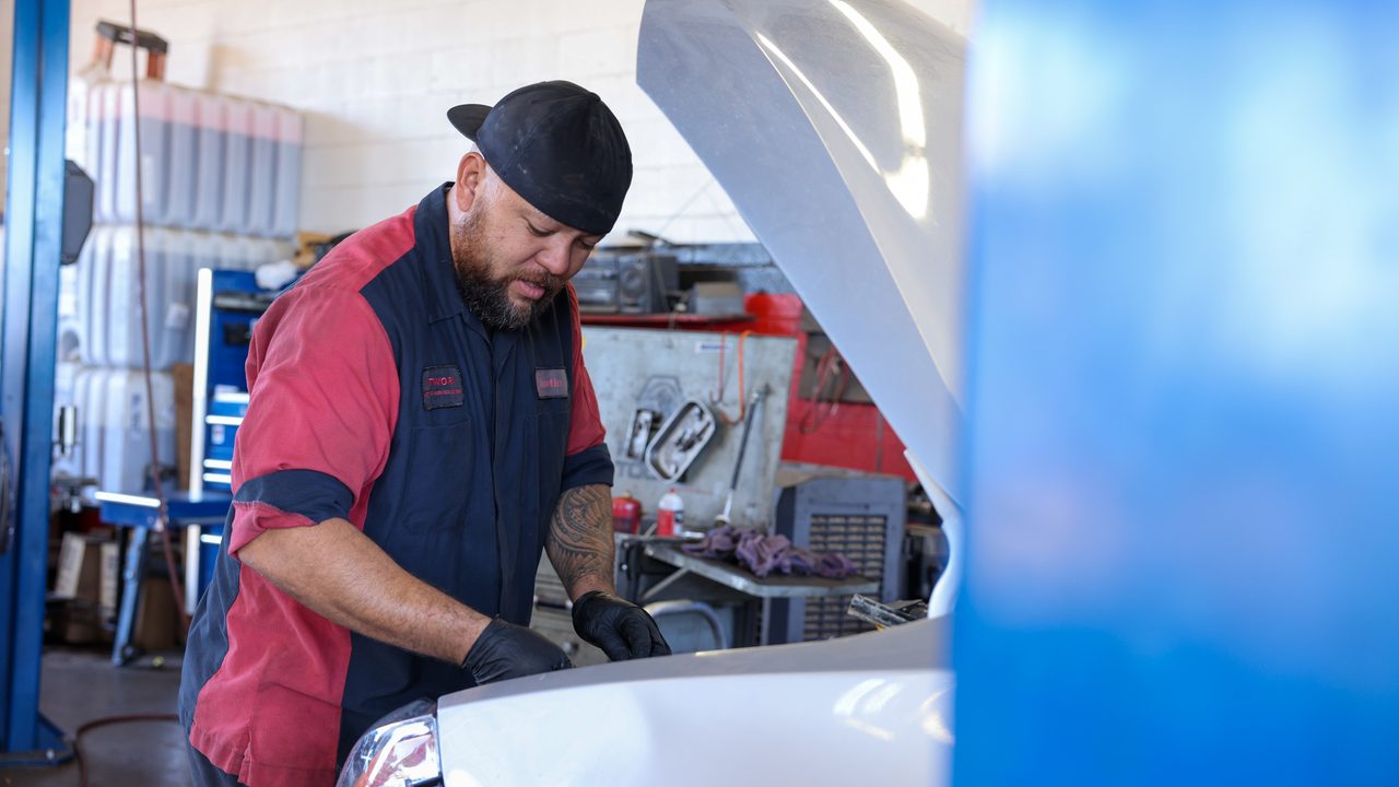 A technician works under the open hood of a vehicle inside an auto repair service bay