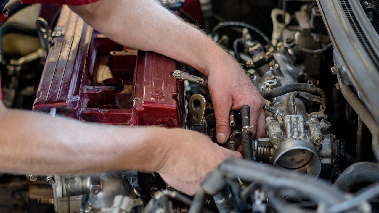 A technician works with both hands on engine components inside the vehicle's engine bay