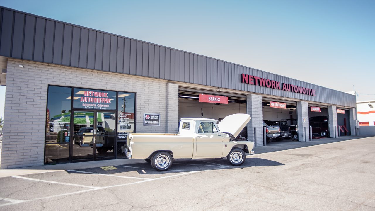 A classic pickup truck with its hood open sits parked outside an automotive repair shop's service bays