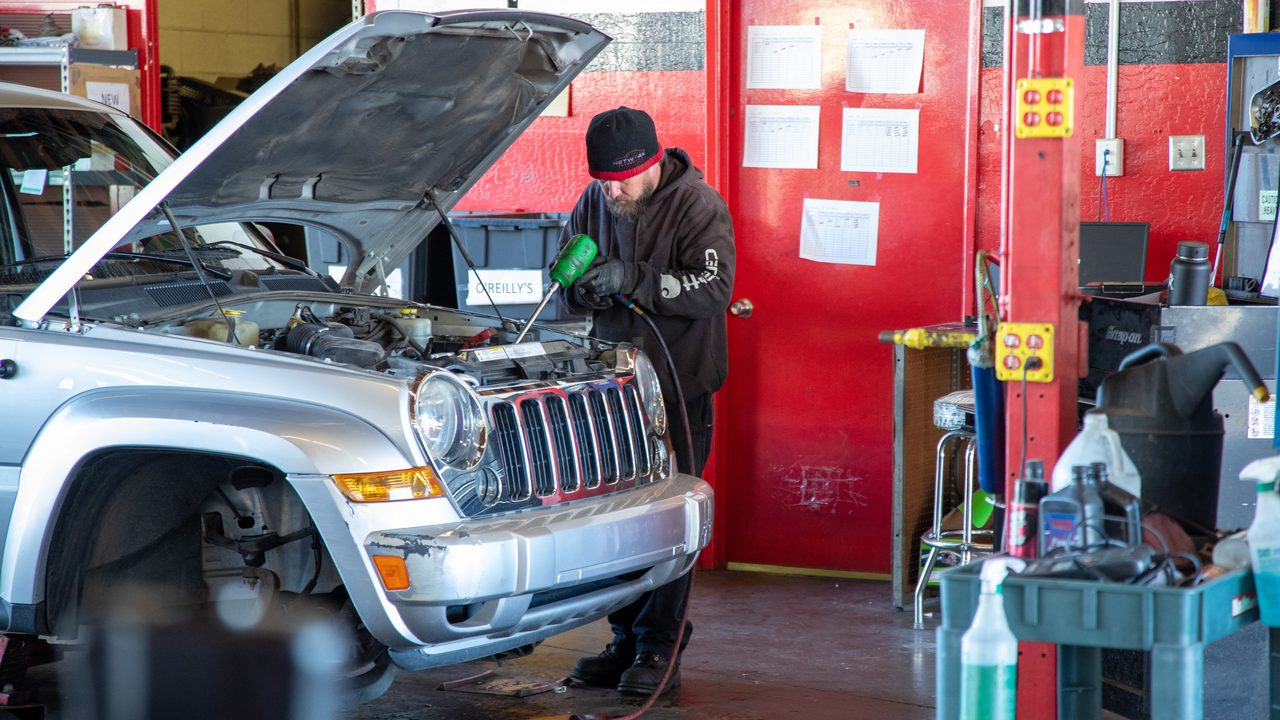 A technician works on the open engine bay of a silver SUV inside an auto repair shop
