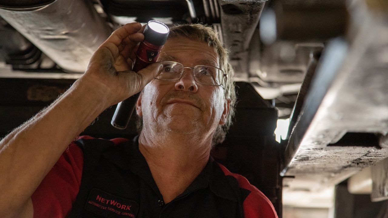 A technician uses a flashlight to inspect the undercarriage of a vehicle raised on a lift