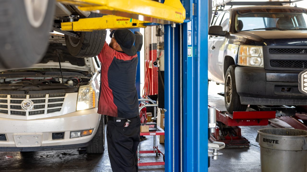 A technician inspects underneath an SUV raised on a lift inside a busy service bay