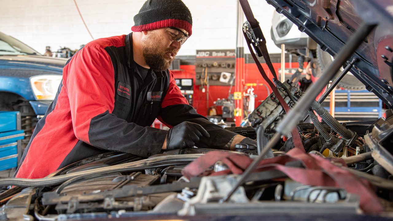 A technician wearing gloves inspects and works on components inside an open car engine bay