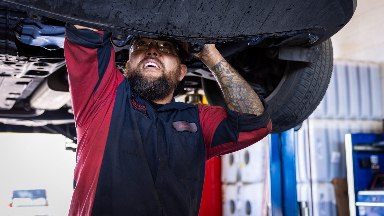 A technician wearing safety glasses works underneath the undercarriage of a vehicle raised on a lift