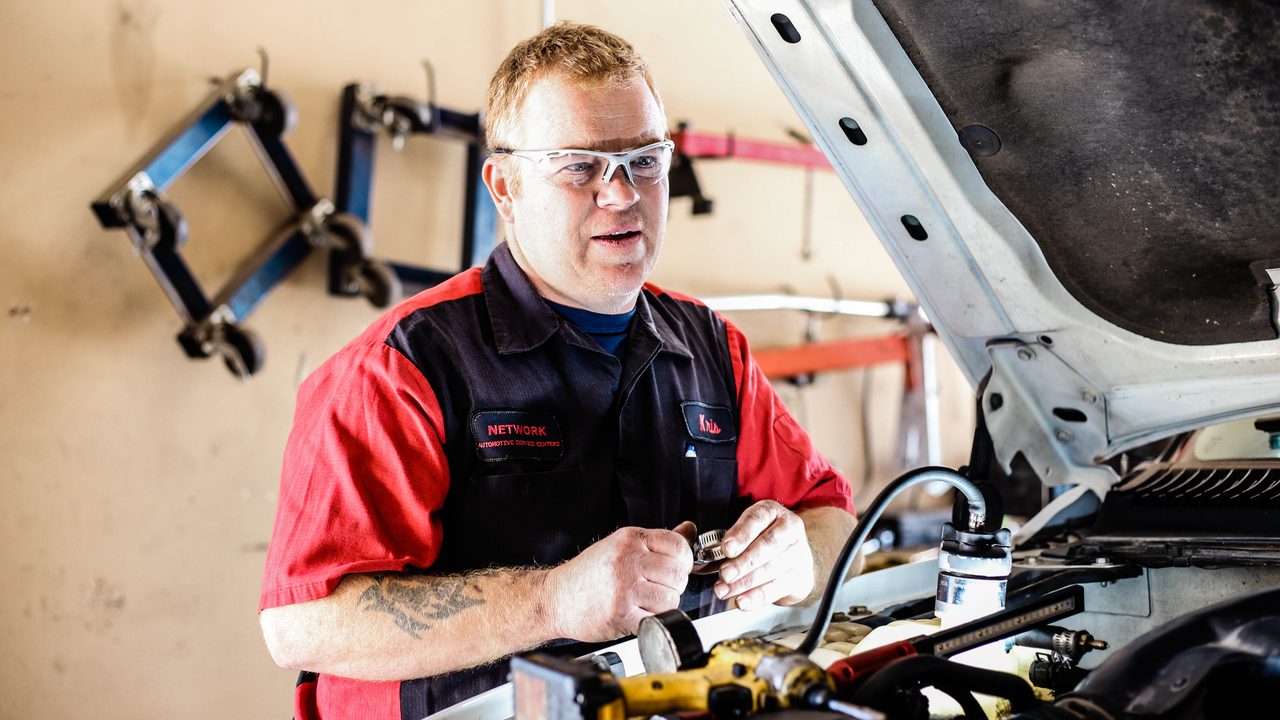 A technician wearing safety glasses works under the open hood of a truck inside a service bay
