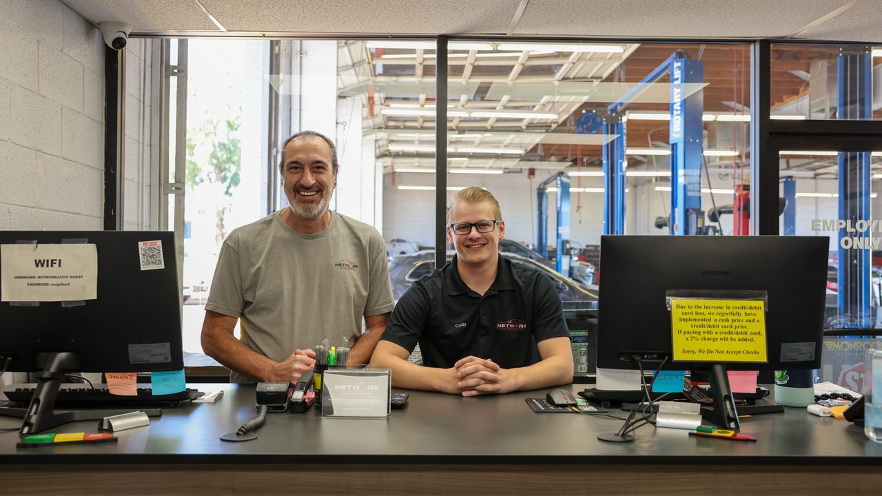 Two auto repair shop staff members smile behind the front counter with the service bay visible behind them