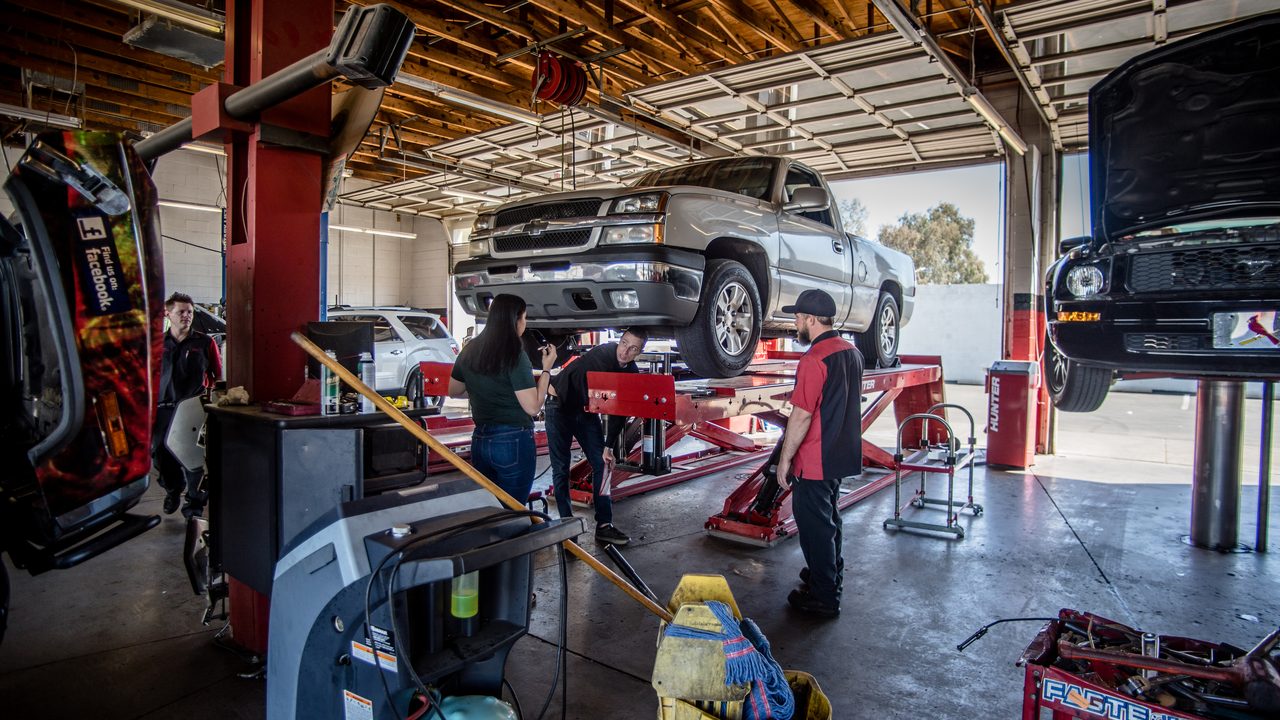 Technicians and a customer inspect a pickup truck raised on a scissor lift inside a busy service bay