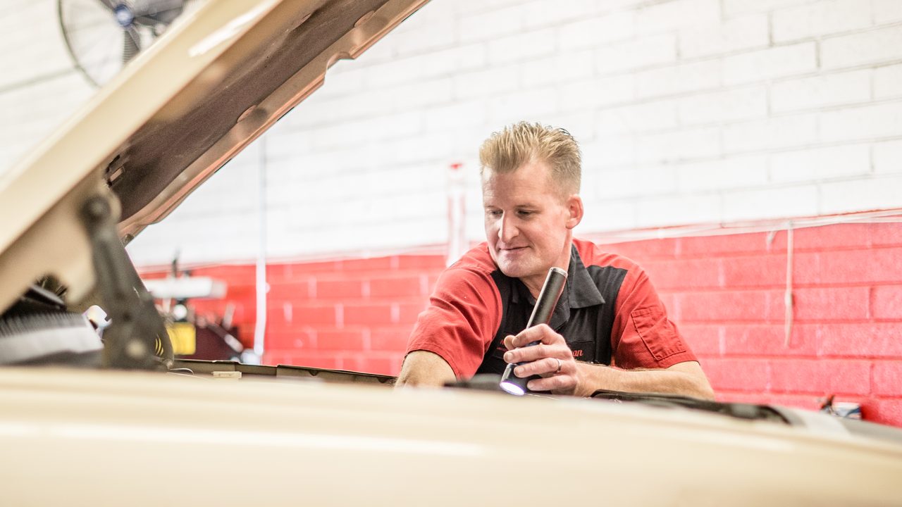 A technician uses a flashlight to inspect components in an engine bay under an open hood