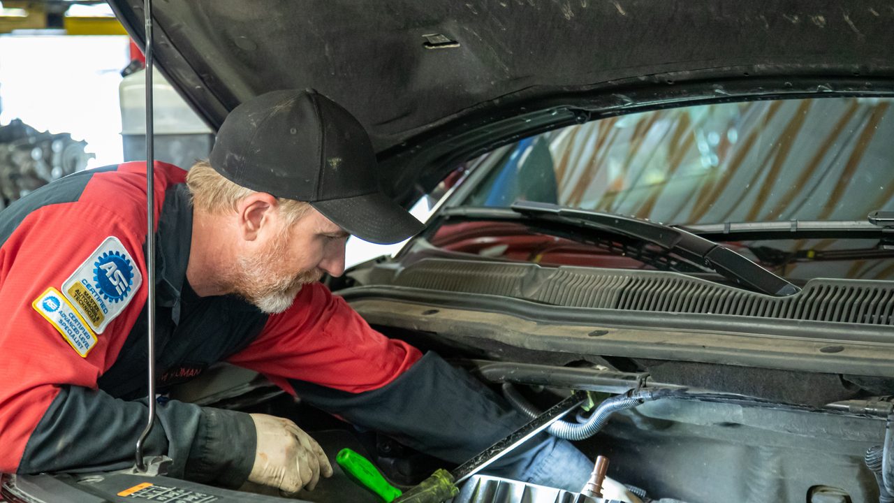An ASE-certified technician inspects and repairs components inside an open vehicle engine bay