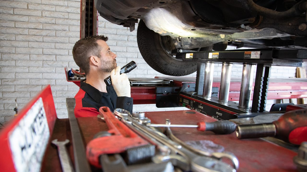 A technician uses a flashlight to inspect the undercarriage of a vehicle raised on a lift