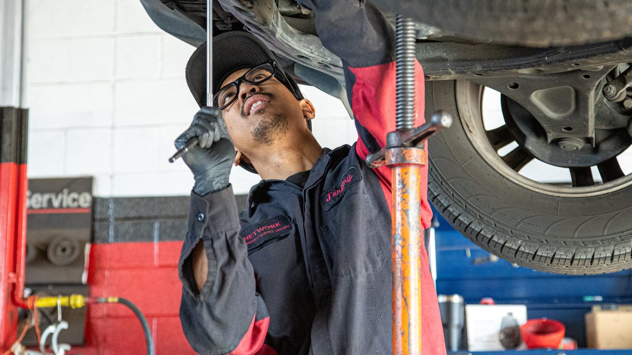 A technician uses a hand tool to work on the undercarriage of a vehicle on a lift