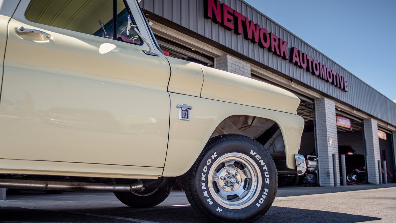 A classic pickup truck is parked outside an automotive repair shop with open service bays