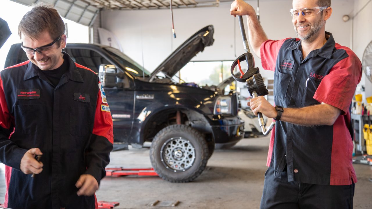 Two technicians smile while handling equipment in an auto service bay with a lifted pickup truck