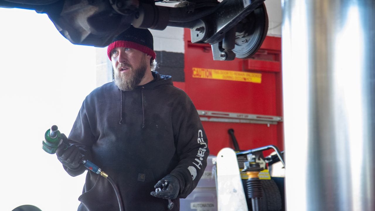 A technician holds tools while working beneath a vehicle raised on an automotive lift in a service bay