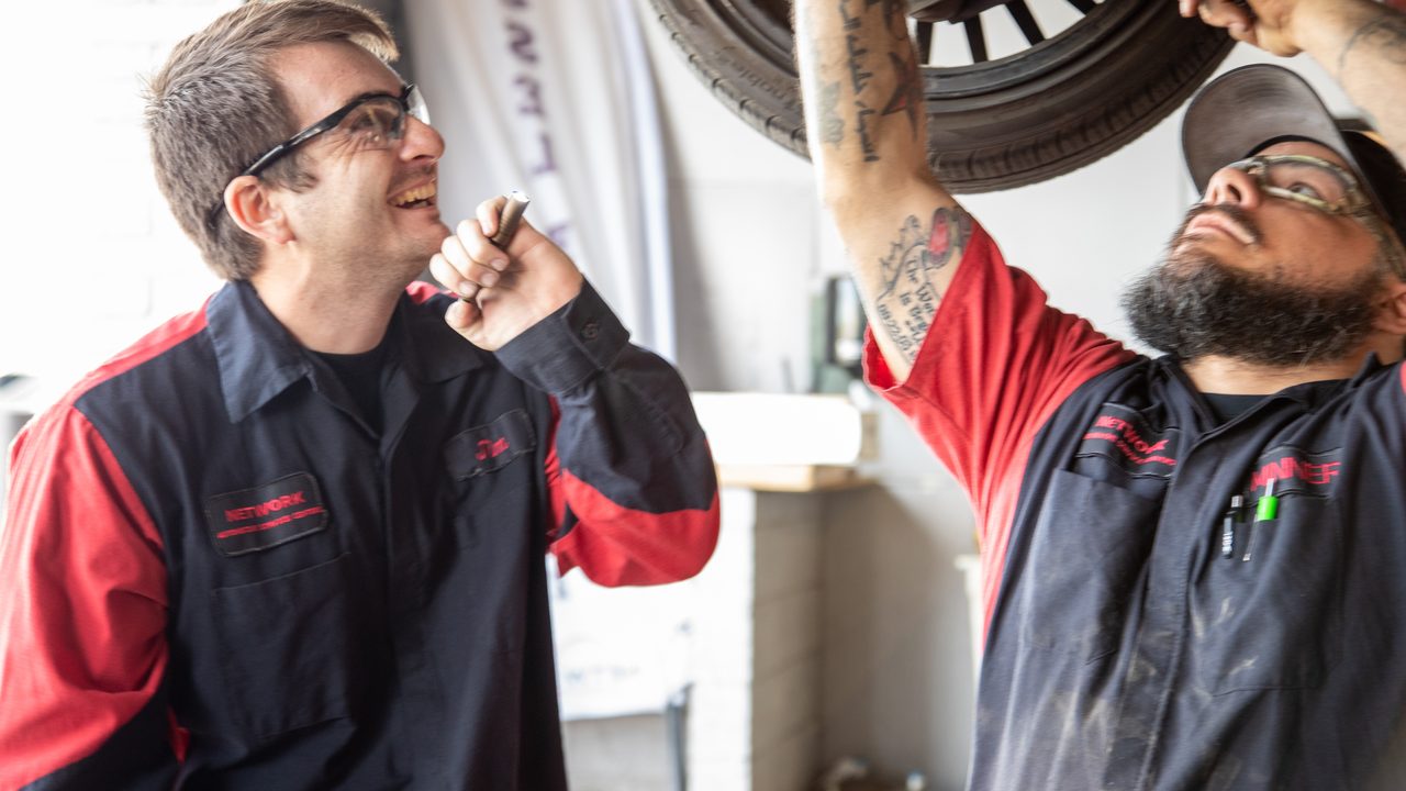 Two technicians inspect a vehicle's suspension and undercarriage while it is raised on a lift