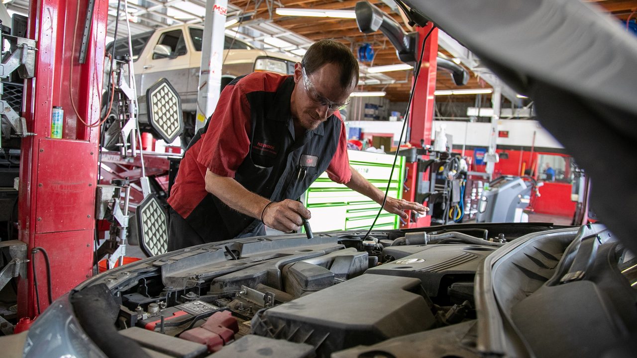 A technician wearing safety glasses inspects a car's engine bay inside an auto repair shop