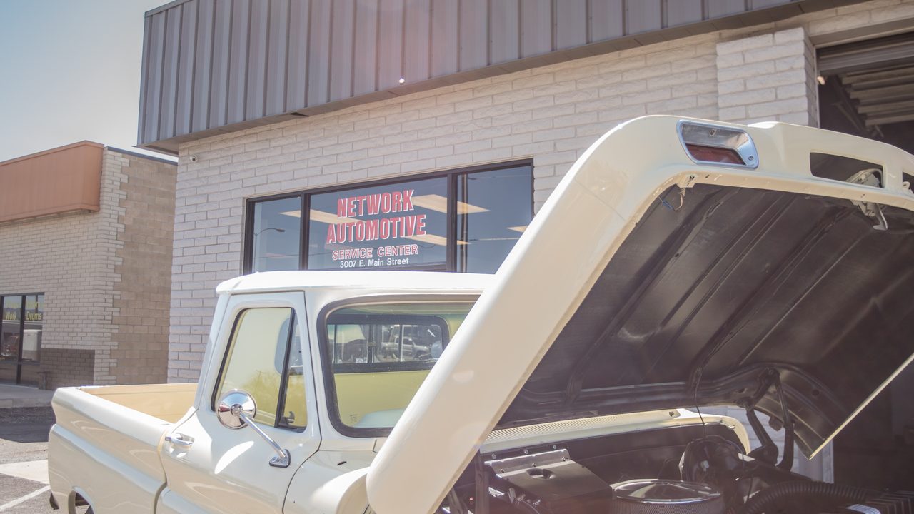 A classic pickup truck sits parked outside an auto repair shop with its hood raised, exposing the engine bay