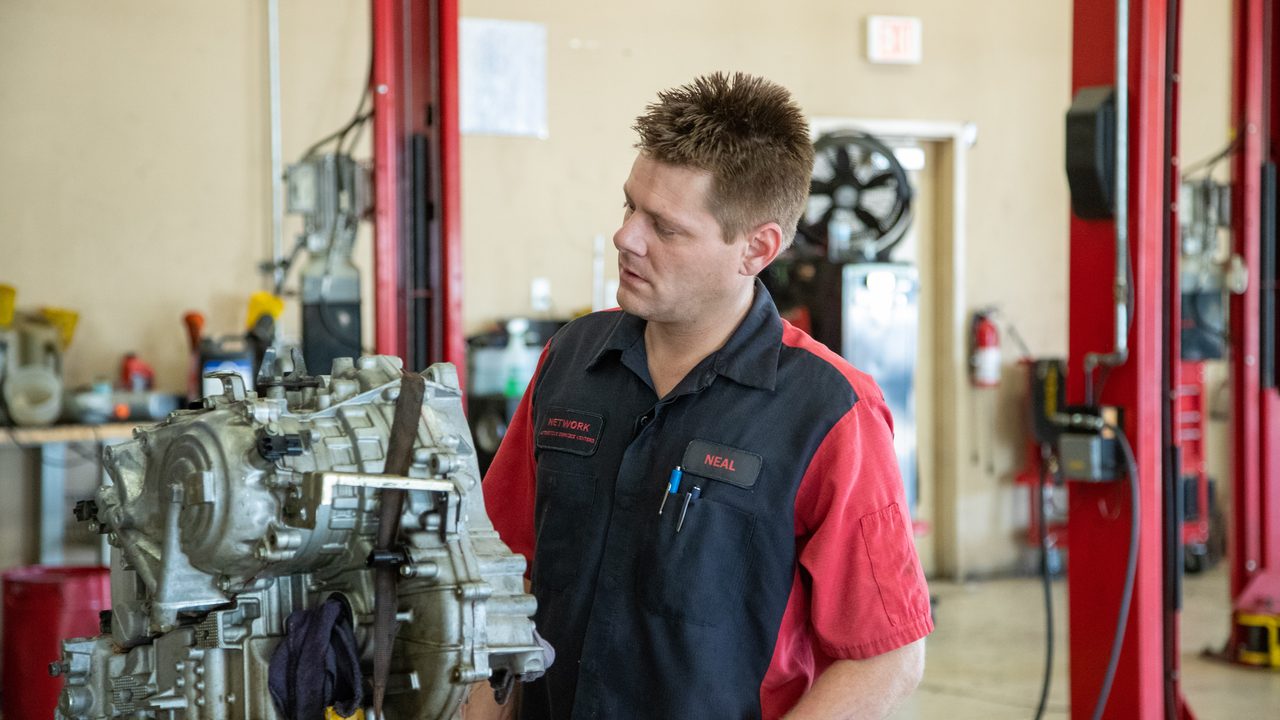 A technician inspects a removed transmission strapped to a jack inside an auto repair service bay