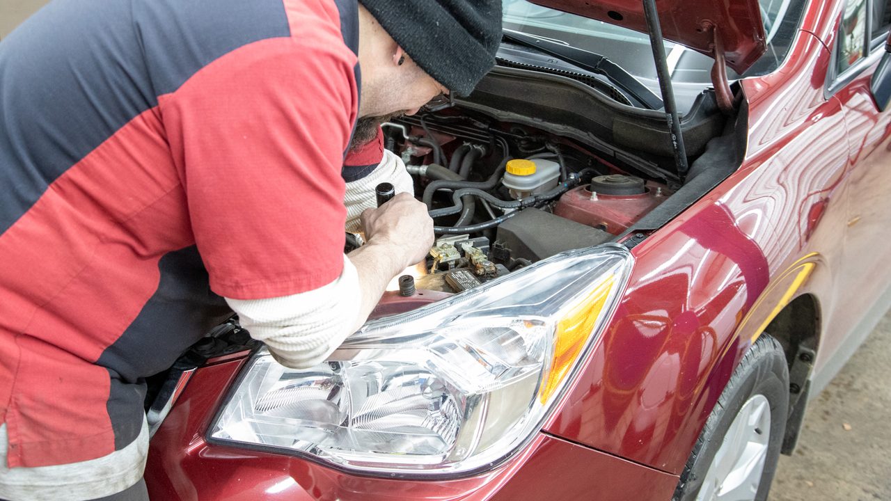 A technician uses a flashlight to inspect components inside the engine bay of a red car