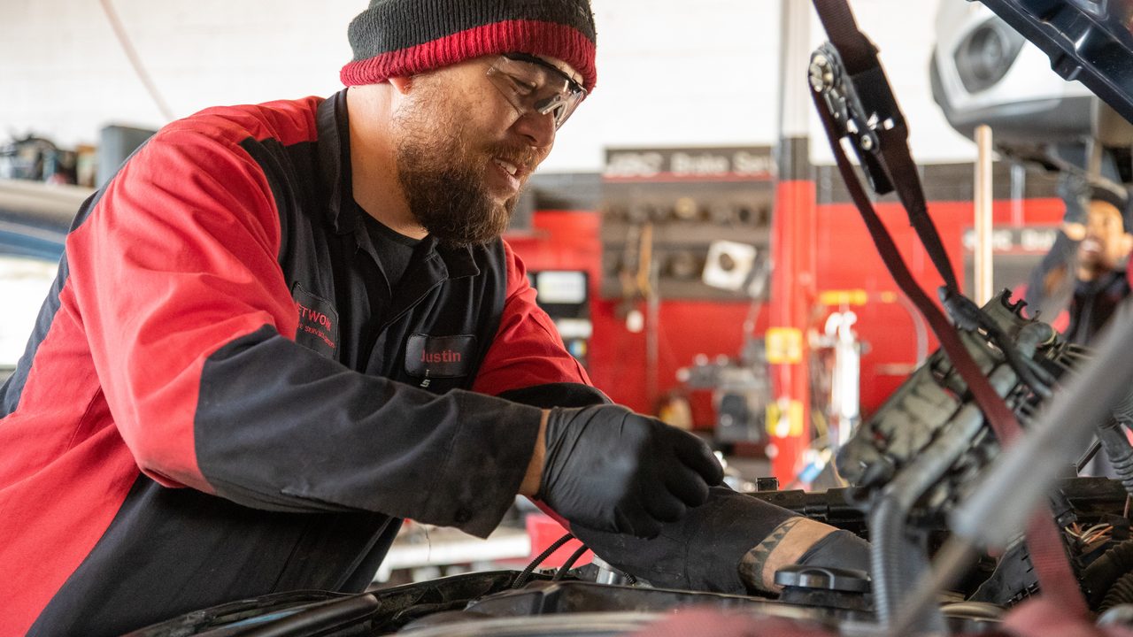 A technician inspects and works on engine components inside an open engine bay in an auto repair shop