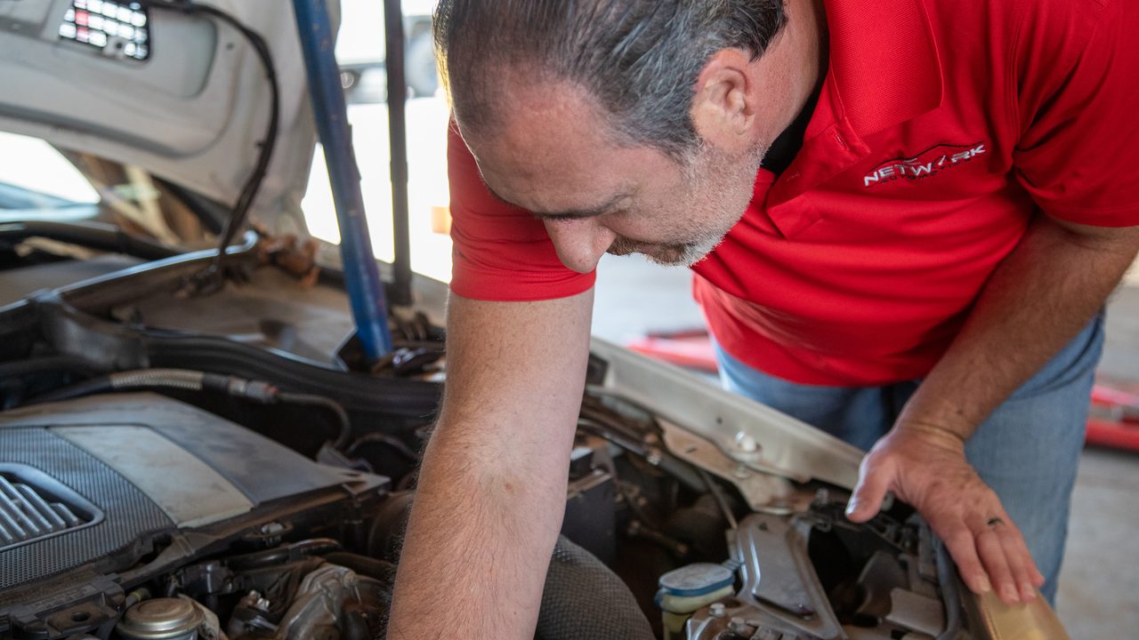 A technician leans over an open vehicle hood to inspect components inside the engine bay