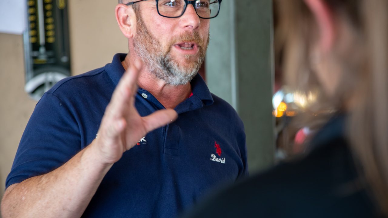 A technician wearing glasses gestures as he explains vehicle service details to a customer