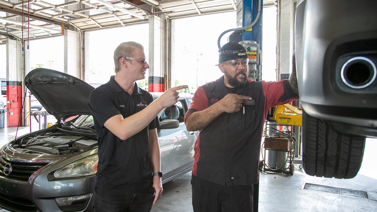 Two technicians discuss vehicle repairs beside a car raised on a lift in an auto shop