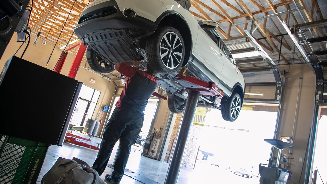 A technician works underneath a white SUV elevated on a hydraulic lift inside a service bay