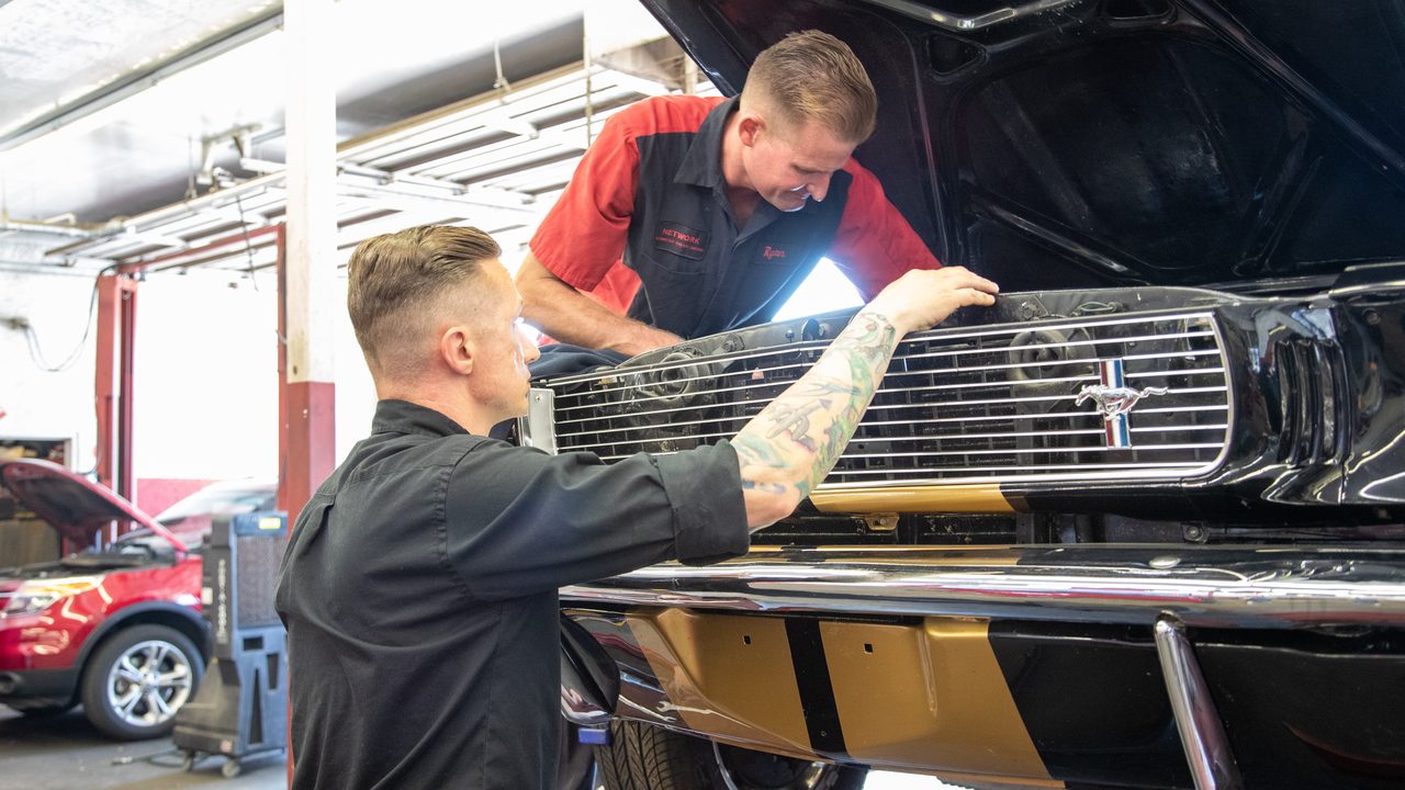 Two technicians inspect under the open hood of a classic car in an auto repair service bay