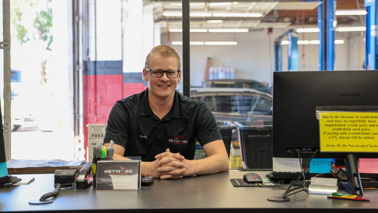 A service advisor sits at the front counter of an auto repair shop with the service bay behind him