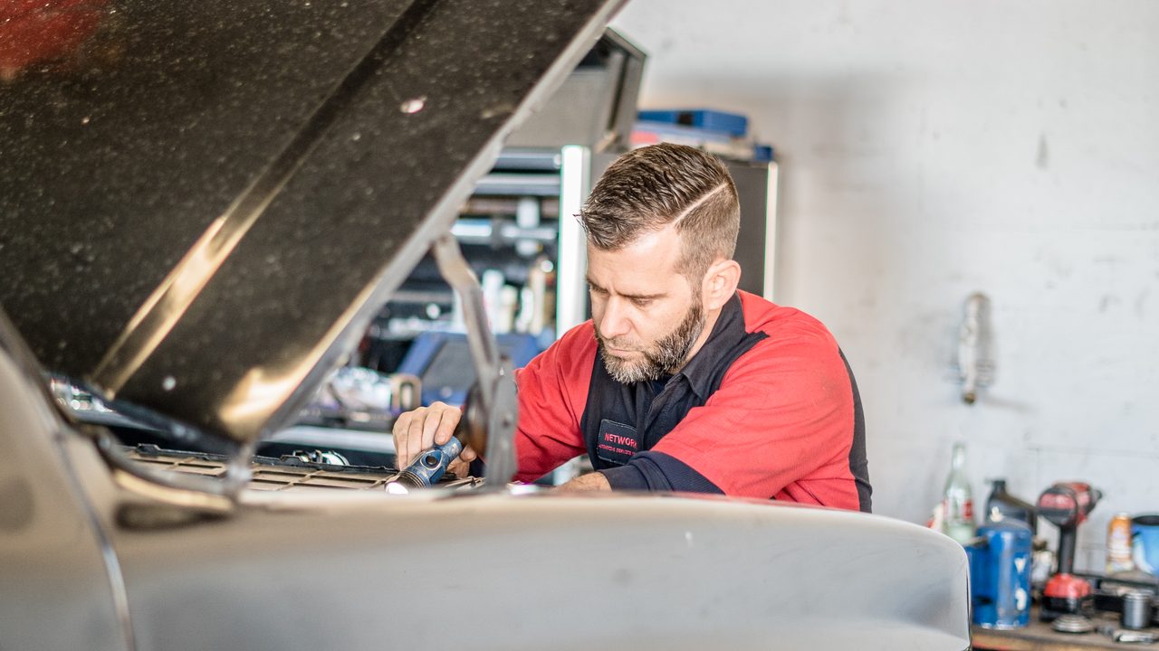 A technician uses a work light while inspecting components inside the open engine bay of a vehicle