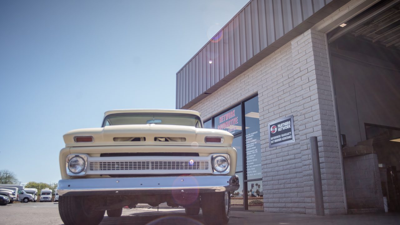 A vintage pickup truck is parked outside an auto repair shop storefront and open service bay